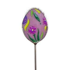 May include: A pink egg-shaped pin with a floral design in shades of green, purple, and yellow. The pin is attached to a silver metal stem.