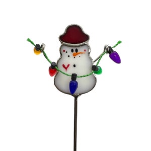 May include: A white snowman figurine with a red hat and a string of colorful lights. The snowman is holding the lights in its arms.