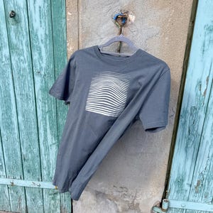 Könnte beinhalten: Ein graues T-Shirt mit einem weißen, wellenförmigen quadratischen Design. Das kurzärmelige Shirt hängt an einem grauen Kleiderbügel vor einer verwitterten, türkis bemalten Holztür und einer Betonmauer.