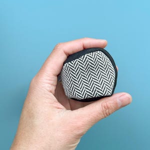 Könnte beinhalten: Ein kleines, quadratisches Objekt mit einem schwarz-weißen Fischgrätmuster wird in einer Hand vor blauem Hintergrund gehalten. Das Objekt hat einen schwarzen Rand. Das Gesamtdesign ist einfach und geometrisch.