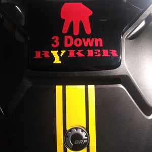 Peut inclure: Composant de v&eacute;hicule noir avec un symbole de main rouge et le texte "3 Down RYKER" en rouge et jaune. Deux bandes jaunes descendent au centre. Le logo BRP est en bas.
