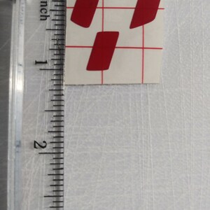 May include: Three red vinyl stickers in the shape of a slightly rounded rectangle. The stickers are on a white sheet of paper with a grid pattern.