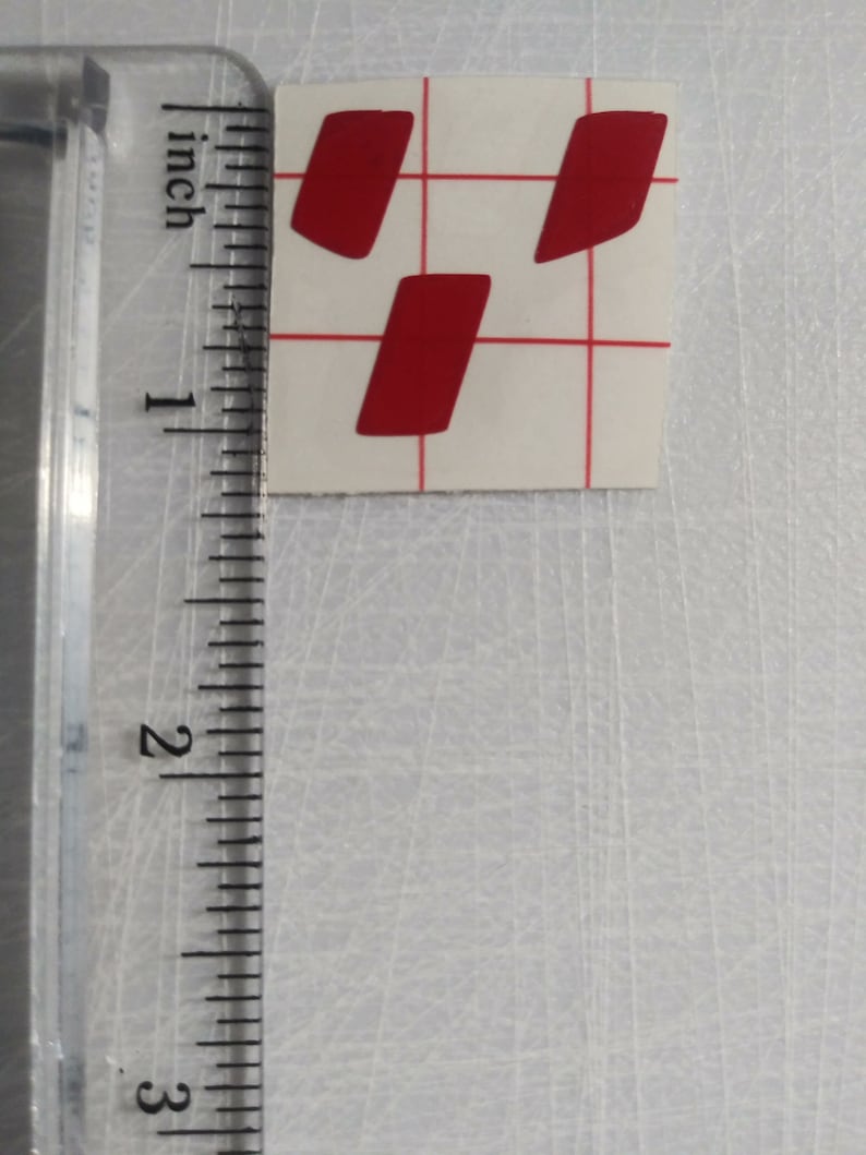 May include: Three red vinyl stickers in the shape of a slightly rounded rectangle. The stickers are on a white sheet of paper with a grid pattern.