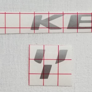 May include: A silver vinyl sticker with the word "RKEIR" in a bold, stylized font. The sticker is on a white background with a grid pattern.