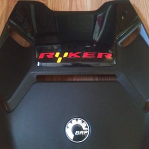 May include: Black motorcycle seat cover with a red and yellow "R&Uuml;KER" logo and a white and gray BRP logo.