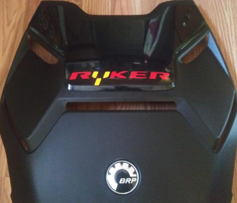 May include: Black motorcycle seat cover with a red and yellow "R&Uuml;KER" logo and a white and gray BRP logo.