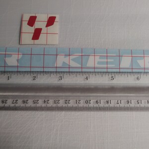 May include: White vinyl sticker with the word "TRIKER" in white letters on a blue grid background. The sticker is measured with a ruler in inches.