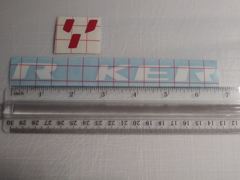 May include: White vinyl sticker with the word "TRIKER" in white letters on a blue grid background. The sticker is measured with a ruler in inches.