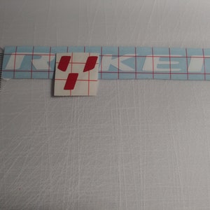 May include: White vinyl sticker with the word "TRUCKER" in white letters. The "R" in "TRUCKER" is highlighted in red.