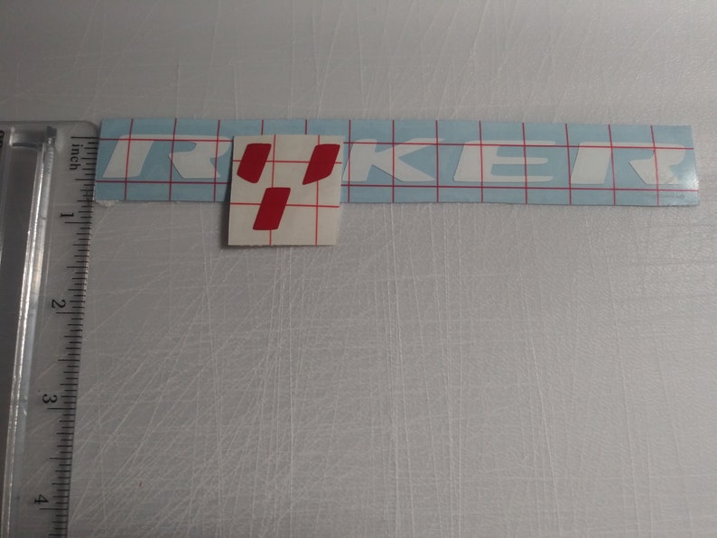 May include: White vinyl sticker with the word "TRUCKER" in white letters. The "R" in "TRUCKER" is highlighted in red.