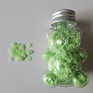 May include: A clear, bear-shaped bottle filled with light green star-shaped confetti. The bottle has a silver cap. A small pile of matching confetti is next to the bottle on a white surface.