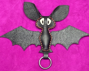 May include: A black leather bat keychain with red stitching and a cute cartoon face. The bat has large, rounded ears and a small, round nose. The keychain has a silver ring.