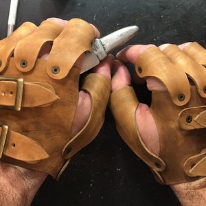 May include: A pair of brown leather fingerless gloves with buckles and rivets. The gloves are worn on a person's hands.