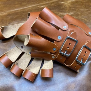 May include: Brown leather gloves with multiple finger slots and two adjustable buckles. The gloves are made of thick, durable leather and have a rustic look.