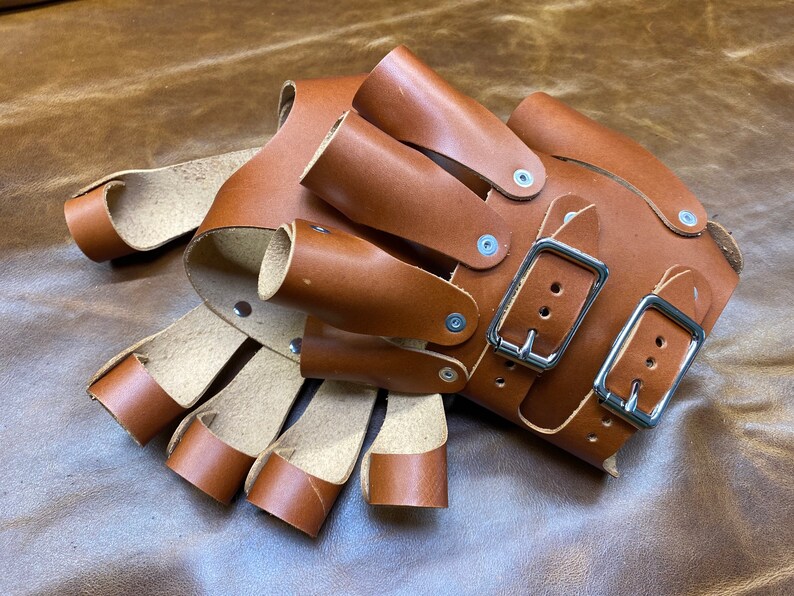 May include: Brown leather gloves with multiple finger slots and two adjustable buckles. The gloves are made of thick, durable leather and have a rustic look.