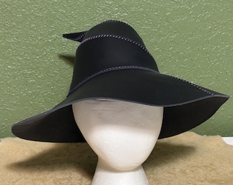 May include: A black witch hat with a wide brim and a pointed crown. The hat features decorative purple stitching along the seams and edges. The hat is displayed on a white foam head.