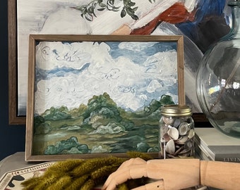 May include: A framed painting of a landscape with a cloudy sky, green trees, and a field. The painting is on a table with a wooden hand, a jar of seashells, and a book titled "Living Again".