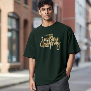 May include: Dark green t-shirt with the text "just stop and pray" in a gold and white script. The shirt is a solid color and has short sleeves. The person is wearing gray pants.