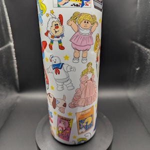 May include: A white tumbler with a colorful design featuring cartoon characters and retro game graphics. The design includes characters like Care Bears, Cabbage Patch Kids, and a Ghostbusters character. The tumbler has a blue lid and a black base.