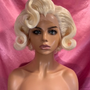 May include: A platinum blonde wig with voluminous, deep-set curls styled in a classic shoulder-length bob. The wig features a sheer lace front hairline, visible lace mesh, and a side-parted design, displayed on a smooth, neutral-toned mannequin head.