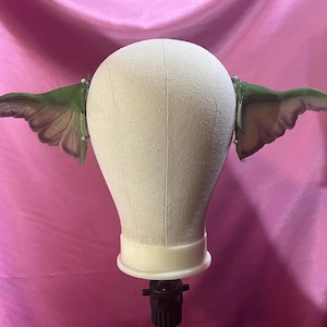 May include: A pair of green and brown fairy wings attached to a mannequin head. The wings are detailed with veining and gradient colors, and are attached with small clips. The mannequin head is a neutral beige color, set against a pink backdrop.