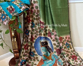 May include: A green and cream colored silk saree with a floral design and a painted image of a deity playing a flute. The saree has a green border with small beads and a blue blouse with floral embroidery.