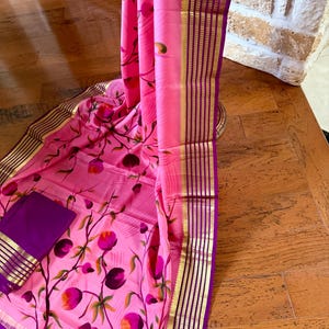 May include: A pink floral printed fabric with a purple border and gold accents. The fabric features a repeating pattern of stylized flowers in shades of pink, purple, and green. A matching solid purple fabric is also included.