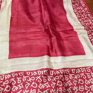 Pode incluir: Um lenço de seda vermelho com uma borda branca que apresenta um padrão repetitivo de escrita branca. O lenço está dobrado ao meio e colocado sobre uma superfície de madeira.