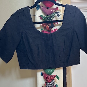 May include: A black, short-sleeved blouse with a round neckline and eyelet detailing. The blouse is on a hanger and has a tie closure at the front.