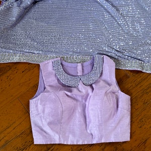 Pode incluir: Uma blusa sem mangas lavanda com uma gola de lantejoulas. A blusa tem um corpete justo e uma bainha reta.