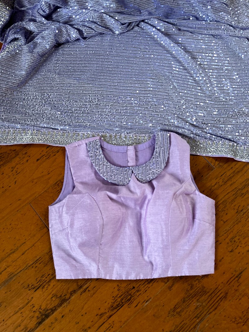 Pode incluir: Uma blusa sem mangas lavanda com uma gola de lantejoulas. A blusa tem um corpete justo e uma bainha reta.