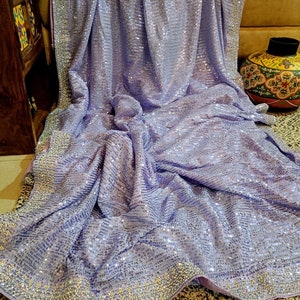 Pode incluir: Um saree de cor lavanda com um design de lantejoulas e uma borda de contas prateadas. O saree est&aacute; drapeado sobre um sof&aacute; e tem uma textura macia e fluida.