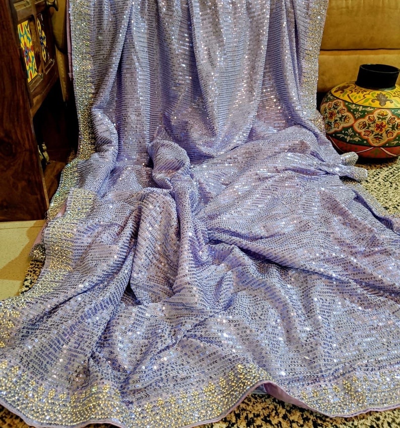 Pode incluir: Um saree de cor lavanda com um design de lantejoulas e uma borda de contas prateadas. O saree est&aacute; drapeado sobre um sof&aacute; e tem uma textura macia e fluida.
