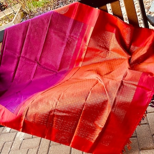 May include: A vibrant textile with a gradient of colors, transitioning from deep magenta to fiery orange and red. The fabric is draped over a wooden bench, showcasing its intricate weave and rich hues. The edges are trimmed with a decorative border.