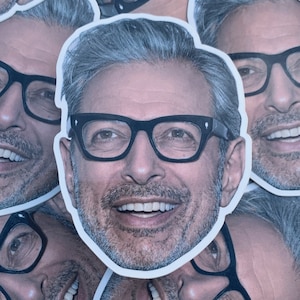 May include: A close-up of a sticker featuring a smiling man with gray hair and glasses. The sticker is printed on white paper and has a rounded shape.