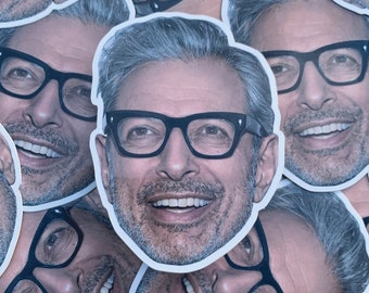 Puede incluir: Primer plano de una pegatina con un hombre sonriente con el pelo gris y gafas. La pegatina está impresa en papel blanco y tiene una forma redondeada.