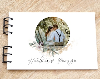 May include: A white guest book with a black spiral binding. The cover features a circular photo of a couple in a forest setting, surrounded by watercolor floral details. The text "Heather & George" is written in cursive below the photo.