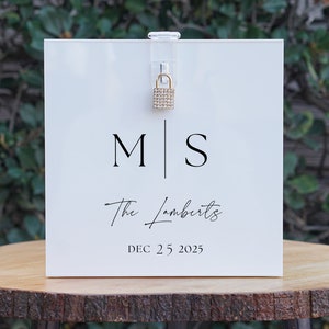 May include: White acrylic box with a gold padlock and a sign that reads "M|S The Lamberts DEC. 25 2025".