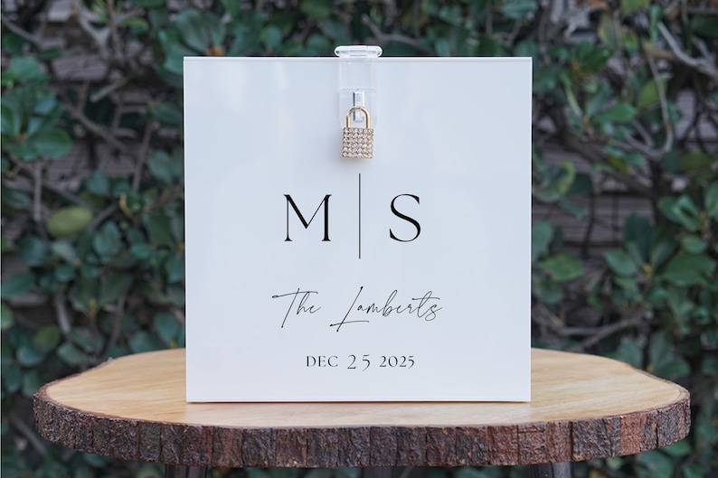 May include: White acrylic box with a gold padlock and a sign that reads "M|S The Lamberts DEC. 25 2025".