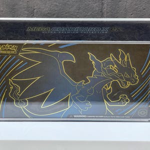 May include: A clear acrylic display case containing a Mega Charizard X EX Ultra-Premium Collection box. The box features a black and gold design with a Charizard illustration and the Pokémon Trading Card Game logo. The case sits on a white surface.