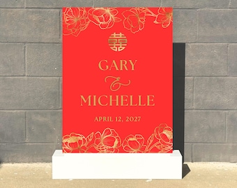 May include: A red wedding sign with gold floral accents and text. The sign displays the names "Gary & Michelle" and the date "April 12, 2027". The sign is placed on a white rectangular base, with a brick wall in the background.