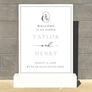 May include: A white wedding welcome sign with a green border, standing on a white base. The sign features the names "Taylor and Henry" and the text "Welcome to our wedding". The date "August 15, 20XX" is also displayed.