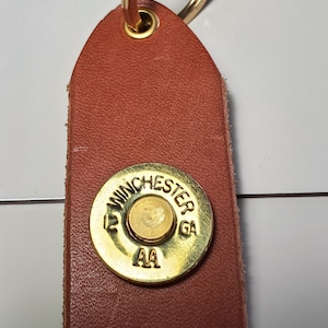 May include: A brown leather keychain with a gold-colored Winchester 12 GA AA shotgun shell attached to a gold-colored key ring.
