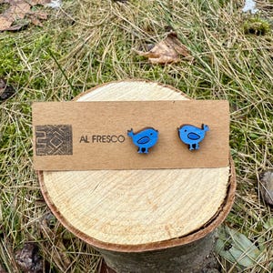 May include: A pair of blue bird-shaped earrings on a wooden display card. The earrings are made of wood and painted blue with black details. The display card has the text "AL FRESCO" and a logo. The earrings are displayed on a small wooden slice.