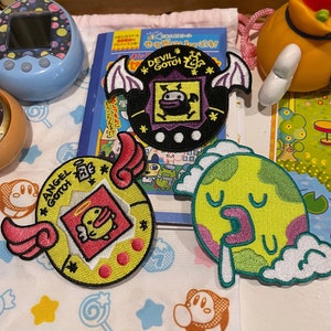 Puede incluir: Parches bordados con diseños de dibujos animados. Un parche tiene un diseño de diablo con el texto "DEVIL GOTCH". Otro parche tiene un diseño de ángel con el texto "ANGEL GOTCH". Un tercer parche es un planeta con una cara. Los parches están sobre una superficie estampada.