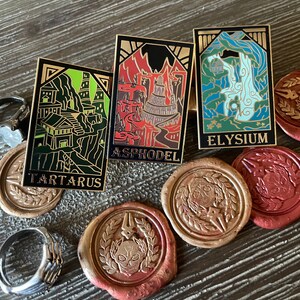 May include: Three rectangular enamel pins with gold borders, featuring detailed illustrations and the names Tartarus, Asphodel, and Elysium. The pins are set on a wooden surface with decorative wax seals and a silver ring.