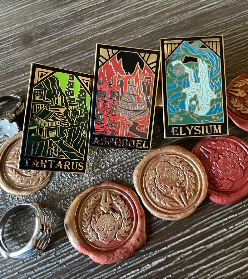 May include: Three rectangular enamel pins with gold borders, featuring detailed illustrations and the names Tartarus, Asphodel, and Elysium. The pins are set on a wooden surface with decorative wax seals and a silver ring.