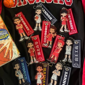 Puede incluir: Una colección de llaveros y etiquetas de personajes de baloncesto estilo anime. Los llaveros presentan varios jugadores con uniformes rojos, blancos y azules. La palabra "SHOHOKU" está bordada en una chaqueta negra. Una pequeña pelota de baloncesto descansa sobre la chaqueta.