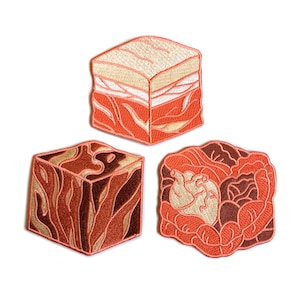 Op de afbeelding: Drie geborduurde patches in tinten oranje, bruin en beige. Eén patch is een kubus met gelaagde secties, een andere is een kubus met een marmer effect, en de derde is een gestileerd hart in een bloemmotief.