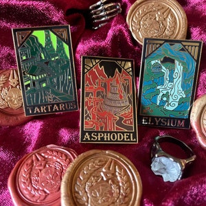 May include: Enamel pins with gold trim and the words "TARTARUS", "ASPHODEL", and "ELYSIUM". The pins are on a burgundy velvet surface, along with gold wax seals and a skeleton hand ring.